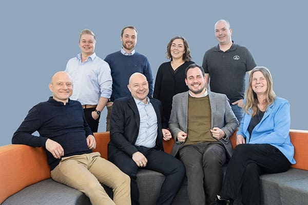 Hydrogen Valley - Fotograf Henrik Espensen Professionelt gruppefoto på blå baggrund for Hydrogen Valley af medarbejdere siddende på og stående bag en rund modulsofa med orange ryglæn og grå sæde fotograferet af fotograf i Randers Fotograf Henrik Espensen